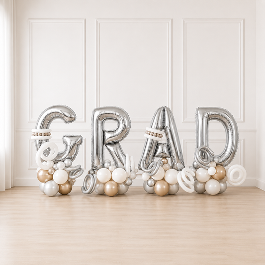 Signature GRAD balloon letter display featuring oversized silver foil balloon letters with white, silver, and champagne balloon clusters. Luxury graduation balloon décor for high school, college, and university graduation parties.